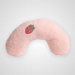A light pink, fuzzy U-shaped neck pillow featuring a small embroidered red strawberry with green leaves on the front, set against a plain light gray background.