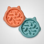 Two round cat bowls with ear cutouts, shown on a light gray background. One bowl is peach-orange and the other is teal-blue, both with raised textured ridges inside.