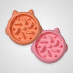 Two round cat bowls with ear cutouts and textured anti-slip interiors, shown on a light gray background. One bowl is peach-orange and the other is pink.